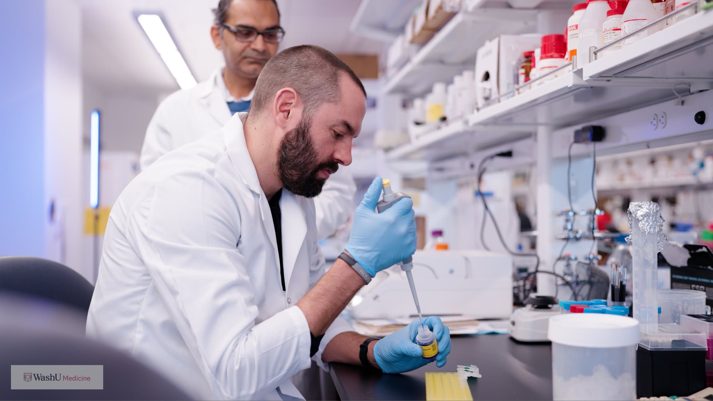A researcher in a white coat and blue gloves pipetting into a small vial at a lab bench while a colleague looks on, in a Washington University Medicine research lab