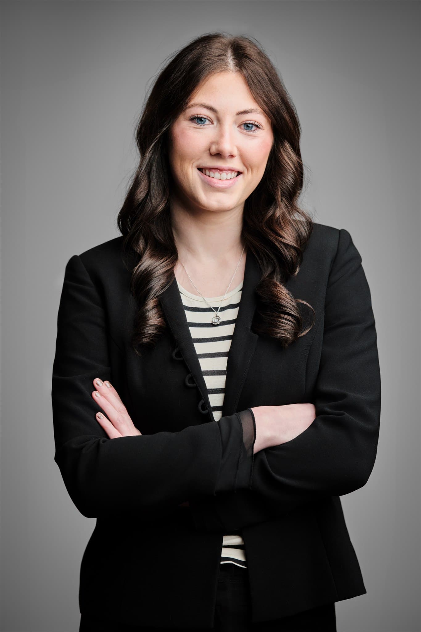 An IWR North America team member in a black blazer over a striped top, arms crossed and smiling, photographed against a grey studio backdrop