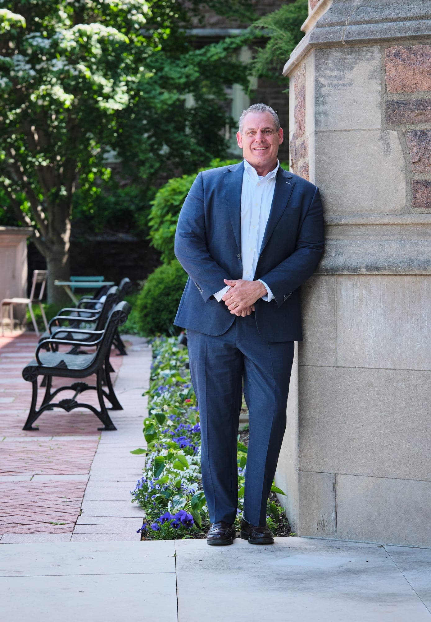 An MHS Legacy executive in a navy suit leaning against a limestone pillar beside a garden path and iron benches at Washington University's Brookings Hall, photographed in natural light